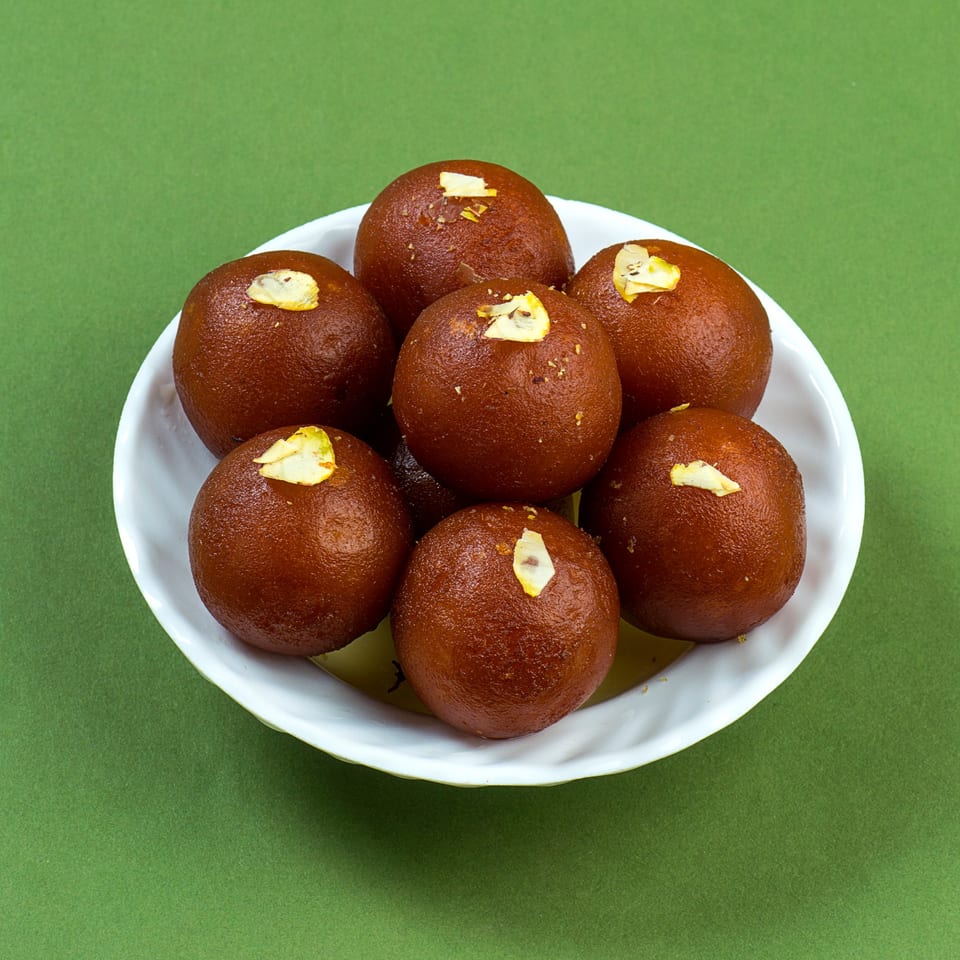 A white bowl filled with several round, brown Gulab Jamun, garnished with slivered almonds, on a green background.
